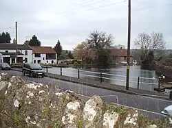 In the foreground are a stone wall and road. Beyond is an area of water surrounded by trees and white fronted houses.