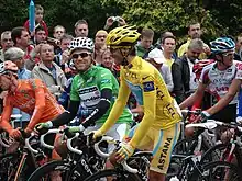 Several cyclists sit on their bicycles. In the center of the image are two, wearing solid yellow and green jerseys. Spectators watch in the background.