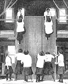 Students co-operate to scale a high wall. The activity involves lifting someone up, wall walking, and pulling someone up; c. 1900.