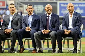 Andy Pettitte, Jorge Posada, Mariano Rivera, and Derek Jeter dressed in suits and seated in chairs on a baseball field.