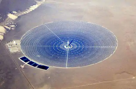 An areal view of a large circle of thousands of bluish mirrors in a tan desert