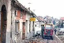 A street of ruined buildings with rubble strewn across the road. A red tractor and other vehicles are visible parked in the background