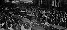 Black-and-white photograph showing a wide and busy street in a city center, with the outer lanes of traffic as well as the sidewalks filled by a massive crowd of people standing in celebration. Only the center two lanes of the road are being used by automobiles. In these center-two lanes is bumper-to-bumper automobile traffic, with the traffic consisting solely of dark-colored personal automobiles of an 1920s-era design. The vehicles are traveling bi-directionally in the North American-style norm of having vehicles drive on the righthand side of their respective direction