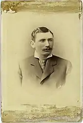 A sepia-toned image of a mustachioed man in a dark Victorian suit