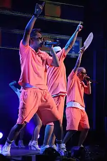 Three men performing on stage with upraised arms, wearing matching neon-orange shorts and polo shirts