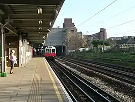 A girl with a pink shirt and blue jeans on a railway platform with a railway track on the right and a white train on it with a red front