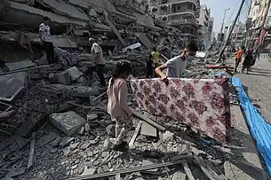 People stand amid the rubble of a building and looking at the ground. A man is carrying a large flower-patterned object.