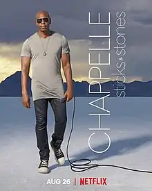 A bald man wearing sunglasses and a long grey tshirt and black pants, stands holding a microphone. He is outside on the plain white ground of the Bonneville Salt Flats and mountains can be seen in the distance.