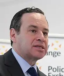 A dark-haired, clean shaven man wearing a jacket and necktie with his mouth slightly open, seen from slightly below. Behind him is a white background on which fragments of words, blurred, can be seen