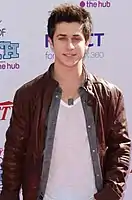 A mid shot of a man with spiked, light brown hair, wearing a brown, leather jacket. He is posing at a press event and smiling, looking directly towards the camera. Behind him are hoarding signs with names of commercial sponsors.