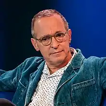 A bust photograph of a white man in spectacles; he is wearing a white patterned shirt, blue jacket, and a jaw-mounted microphone.  He is facing the camera, looking to its left.