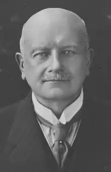 Older male with bald head and chevron moustache wearing a suit, winged collar and tie