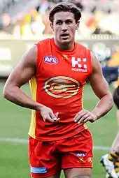 A male athlete with light brown hair wearing a sleeveless jersey and shorts walks on the grass surface of the playing arena.