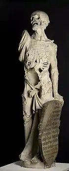 Statue showing Death as a living skeleton. His right arm is missing and he looks upwards, while holding a shield with his right. The shield contains rows of inscriptions.