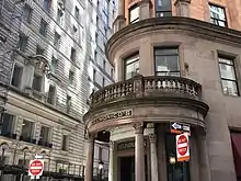 The building's entrance portico, which contains a curved balustrade supported by dark brown columns. There are windows above and next to the portico. A white building is in the background.