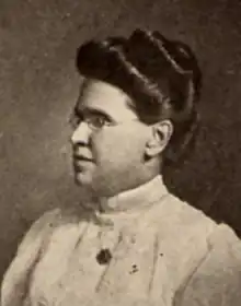 An older white woman, dark hair in a bouffant updo, wearing tiny glasses and a white blouse with a high collar