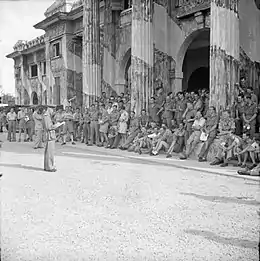 A large group of officers stand in front of a colonnaded building; a senior officer addresses them.