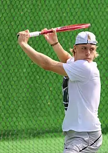 A blonde man with long hair wearing a white shirt, gray shorts, and a backwards white baseball cap holds a red tennis racket in front of his torso and above his head as he looks to his left and prepares to strike