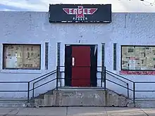 Grey stucco exterior with a red door. Black eagle shaped sign reads "Denver Eagle"