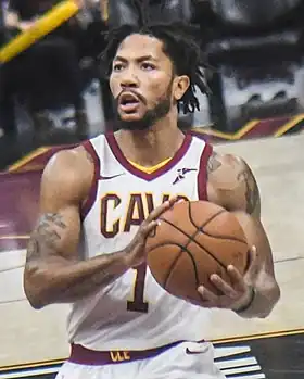 Hip length view of man raising the basketball to shoot free throw, with black moustache, beard, and short dreadlocks, thin white headband, wearing navy blue Timberwolves uniform