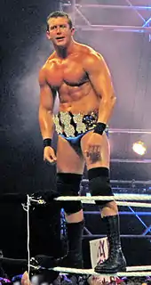 A dark-haired man wearing blue wrestling tights, black knee-high wrestling boots and black kneepads stands on the white ropes of a wrestling ring. He is wearing around his waist a gold-plated professional wrestling championship belt shaped with three dollar signs on the front.