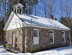 A small stone building viewed from the side in winter. Snow covers the ground and its black roof, which has a small empty steeple and a brick chimney