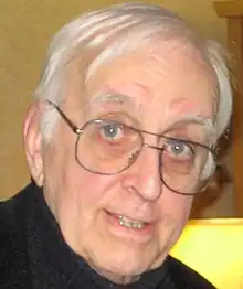 Elderly man with white hair wearing a dark turtleneck sweater and eyeglasses with silver rims