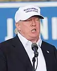 A man in front of a mounted microphone wearing a suit, American lapel pin, and baseball cap