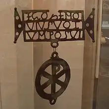 A metal plate with holes. The outline spells 'EGOZENO / VIVSVOT / VM POSVI'. A Chi-Rho shape hangs from it.