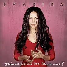 A brunette woman is in front of a pink background. She is wearing a red blouse and colorful braids, and her hands are covered in dirt. The word "Shakira" is above her, stylized in all capital letters, while the words "Dónde Están los Ladrones?" are placed beneath her.