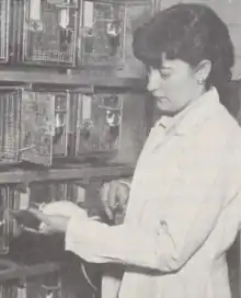 A white woman with dark hair, wearing a laboratory coat and holding a white mouse or rat