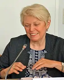 A middle-aged smiling woman with short blond hair wearing a gray sweater over black-and-white patterned top holds her hands flat over a table while sitting in front of a microphone