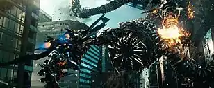 In a city full of skyscrapers, a robot wearing jet thrusters on his back holding a cannon flies toward a large snake-like robot. A tentacle of the larger robot is exploding.