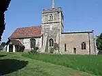 A stone church seen from the south, with a central battlemented tower, the nave with a porch and red tiled roof to the left, and a smaller chancel with a flat copper roof to the right