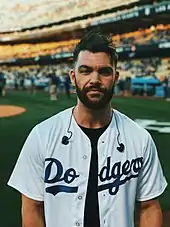 A dark-haired and bearded man standing in a baseball stadium wearing a shirt with "Dodgers" printed on it