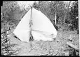 View of a camp site, with a white tent, showing a string of fish and a fly fishing rod hanging from the front tent pole.