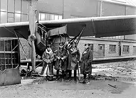 Four men in military uniforms with overcoats, standing next to a biplane parked in front of a building