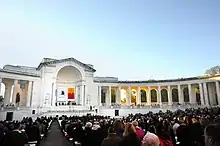 An outdoor auditorium with seated guests lined with neoclassical columns and a closed archway on one side and banners hanging inside the arch.