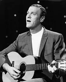 A fair-haired man wearing a turtleneck sweater and a jacket, playing a guitar