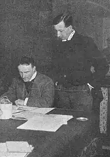 two middle-aged white men, both clean-shaven with full heads of neatly cut dark hair, the one on the left seated, the other standing