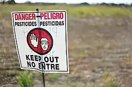 Redd and white caution sign signaling to not enter the designated area behind the sign. In red and black bold letters "DANGER--PELIGRO PESTICIDES--PESTICIDAS KEEP OUT NO ENTRE". An image of a cartoon man holding his hand up signaling a universal sign of stopping.