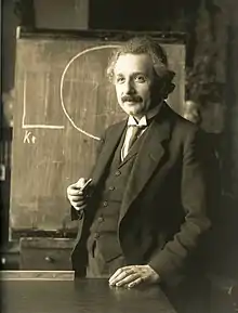 A black-and-white photograph of a man standing in front of a chaulk board, holding a piece of chaulk in his right hand, and resting his left hand on a table