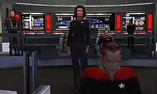 A computer-rendered version of the bridge featured in Star Trek: Voyager. A woman gives orders from the center of the room, while a man works on a console in the foreground. The ship's executive officer sits in a chair to the right, and a crewman works on a panel with the ship's schematics in the background. To the left, a man in an armored uniform watches the activity.