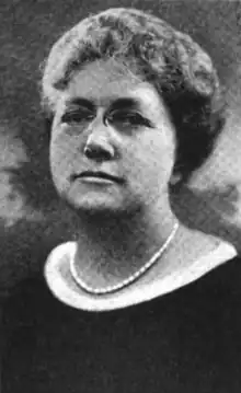 A middle-aged white woman wearing glasses and pearls, and a dark dress with white trim around the neckline