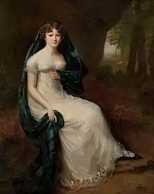 Full length portrait of young woman wearing a white empire-waist dress with a plaid of tartan loosely draped over her head and left shoulder, seated in a park