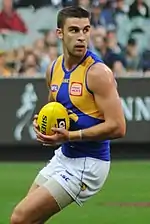 A man with brown hair holds a yellow football on a grassed playing field