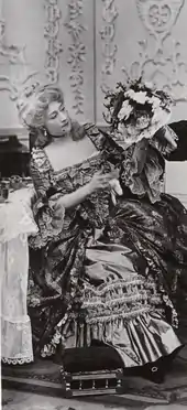 young white woman in elaborate 18th-century costume, seated, looking at a bouquet of flowers