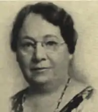 sepiatone headshot image of an older woman with glasses staring at the camera. She is wearing a neacklace