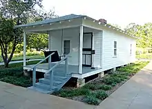 Present-day photograph of a whitewashed house, about 15 feet wide. Four banistered steps in the foreground lead up to a roofed porch that holds a swing wide enough for two. The front of the house has a door and a single-paned window. The visible side of the house, about 30 feet long, has double-paned windows.