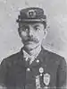 Head and shoulders of a young man with a mustache, wearing a military cap and a jacket with three medals pinned to the left breast.